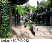 Prague, Czech Republic, July 22, 2018: view of Visegrad cemetery. Tranquil pathway winding through historic cemetery with vibrant green ivy and towering trees. Weathered gravestones and monuments. Редакционное фото, фотограф Olga Monako / Фотобанк Лори