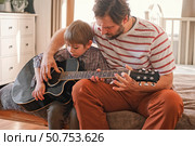 Handsome dad teaches his cute son to play the guitar sitting on sofa in the living room at home. Happy Father's Day. Father teaching his child to play guitar. Kid Learning music skill. Art education. Стоковое фото, фотограф Марина Демидюк / Фотобанк Лори