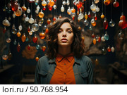 Portrait of a young woman with dark wavy hair, wearing an orange shirt and a denim jacket. In the background, there are many multi-colored drop-shaped pendant lights, creating a warm atmosphere. The woman looks straight into the camera with a slight thoughtfulness. The lighting emphasizes her facial features, creating a deep and expressive image. Стоковое AI-изображение, AI-художник Анна Кривицкая / Фотобанк Лори