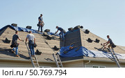 Workers installing roof shingles on a residential house, using ladders, tools, and blue tarp. They are wearing safety harnesses while some carry shingles up to the rooftop. Стоковое AI-изображение, AI-художник Olga Dmitrieva / Фотобанк Лори
