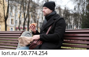 Pensive hipster drinking coffee sitting on a bench in the urban park. Handsome man wearing black coat eating street food, hot dog with paper cup in hand in the city. Stylish bearded middle-aged male. Стоковое видео, видеограф Марина Демидюк / Фотобанк Лори