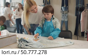 A caring mother is watching her child complete a drawing task at a workshop on creating craft clothing using their own designs. Стоковое видео, видеограф Strunov David / Фотобанк Лори