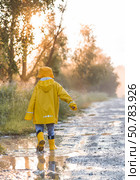 A boy in yellow rubber boots and a raincoat runs through puddles, having fun and playing after it rains. Summer and autumn holidays. A happy childhood. Стоковое фото, фотограф Photojuli / Фотобанк Лори