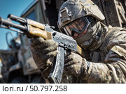 Российский военнослужащий в полной боевой экипировке и защитном шлеме прицеливается из автомата на фоне военной техники в зоне боевых действий. Стоковое AI-изображение, AI-художник ArtVladZ / Фотобанк Лори