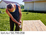 Holding angle grinder with both hands, craftsman wearing respirator and safety glasses sanding tabletop outdoors in rustic countryside setting. Стоковое фото, фотограф Владимир Григорьев / Фотобанк Лори
