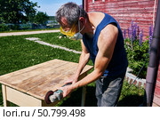 Working with handheld grinder on wooden tabletop, carpenter using protective gear during outdoor sanding in simple rural surroundings. Стоковое фото, фотограф Владимир Григорьев / Фотобанк Лори