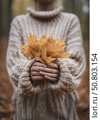 A woman in a cozy knit sweater gently holds a bunch of yellow leaves in a forest, celebrating fall fashion and seasonal warmth. Стоковое AI-изображение, AI-художник Александра Троицкая / Фотобанк Лори