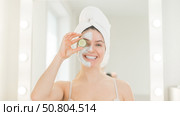 Cheerful woman with a towel on her hair and in a clay face mask fooling around with cucumbers in her hands. Taking care of beauty at home. Стоковое фото, фотограф Михаил Решетников / Фотобанк Лори