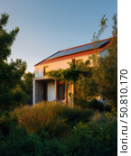 Modern eco house with solar roof panels and lush green vegetation bathed in golden sunset light. Ideal for sustainable living and renewable energy design. Стоковое AI-изображение, AI-художник Александра Троицкая / Фотобанк Лори