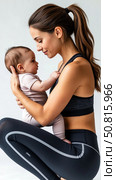 A young mother in a tracksuit holds her newborn baby in her arms. She does fitness exercises with her child. Стоковое AI-изображение, AI-художник Ирина Гильмуллина / Фотобанк Лори