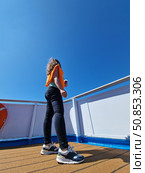 A woman with long hair in an orange T-shirt and black leggings stands on the deck of a ship with a drink in her hand. The bright sun and clear blue sky create a feeling of relaxation and freedom. Стоковое фото, фотограф Анна Чижова / Фотобанк Лори