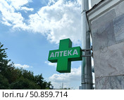 A green pharmacy cross with Cyrillic script "Аптека" (Pharmacy) hangs on a building against a blue sky. Concept of Healthcare access. Стоковое фото, фотограф Ольга Петрова-Апостолова / Фотобанк Лори