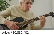 Senior man is playing guitar. Elderly man sitting on the sofa and playing guitar. Portrait of a gray-haired mature man in a sweater and glasses learning to play. Enjoying retirement life at home. Стоковое видео, видеограф Марина Демидюк / Фотобанк Лори
