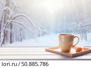Hot beverage placed on windowsill with snowy forest view in winter. Стоковое AI-изображение, AI-художник Иван Рябоконь / Фотобанк Лори