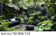 A tranquil garden space enveloped by dense green ferns and broad-leafed plants, with modern seating arranged for relaxation. Стоковое AI-изображение, AI-художник Данила Большаков / Фотобанк Лори