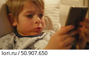 Cute kid boy in pajamas lying in bed watching smartphone, smiling. Children's screen time. Child plays games using phone at the evening or night. Social media addiction. Childhood, modern technology. Стоковое видео, видеограф Марина Демидюк / Фотобанк Лори