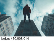 A dramatic low-angle urban shot of a young man in a red hoodie standing on top of a concrete structure, looking up at the sky. To the left and right are tall apartment buildings, adding to the sense of loneliness and immensity. The sky is covered with clouds, creating an atmosphere of reflection, freedom or inner struggle. Ideal for themes of self-determination, urbanism, dark aesthetics, social photography and youth culture. Стоковое AI-изображение, AI-художник Анна Кривицкая / Фотобанк Лори