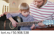 Handsome dad teaches his cute son to play the guitar sitting on sofa in the living room at home. Happy Father's Day. Father teaching his child to play guitar. Kid Learning music skill. Art education. Стоковое видео, видеограф Марина Демидюк / Фотобанк Лори
