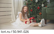 Cute happy little girl with curly hair, in an elegant white fluffy dress sits on floor under decorated Christmas tree at home, joyfully opening a gift box. Childhood and happy family holidays concept. Стоковое видео, видеограф Ирина Михайличенко / Фотобанк Лори