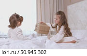 Two children, little sisters sitting on a bed with white bedding, opening gift boxes wrapped in kraft paper. Holidays concept. High quality footage. Стоковое видео, видеограф Ирина Михайличенко / Фотобанк Лори