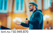 Motivational Speech: Bearded man in hoodie giving presentation with microphone, gesturing with open hands, indoors. Стоковое AI-изображение, AI-художник Данила Большаков / Фотобанк Лори