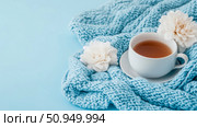 A comforting cup of warm herbal tea rests on a cozy, chunky knitted blue blanket with elegant white blooms, evoking serene winter morning relaxation, ideal for print material. Стоковое AI-изображение, AI-художник Данила Большаков / Фотобанк Лори