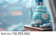 Concept: Tranquil teatime. A steaming ornate turquoise teapot with floral motifs and small cups arranged on an intricately carved wooden table, set against a soft, blurred urban backdrop. Стоковое AI-изображение, AI-художник Данила Большаков / Фотобанк Лори
