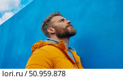 A contemplative man with a beard gazing upwards against a vibrant blue wall and a cloudy sky, representing hope and future aspirations. Стоковое AI-изображение, AI-художник Данила Большаков / Фотобанк Лори