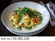 Cauliflower creamy soup with baked cauliflower florets on the top in a white bowl on a dark wood tabletop. Restaurant style dish. Hot, delicious plate of cream-of-cauliflower soup with melted cheese. Стоковое AI-изображение, AI-художник Марина Демидюк / Фотобанк Лори