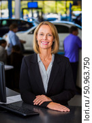 Confident businesswoman in a professional suit stands at a reception desk in a modern automotive dealership, smiling warmly, showcasing customer service and professionalism in a vibrant environment. Стоковое AI-изображение, AI-художник Ирина Ручушкина / Фотобанк Лори