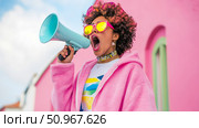 A vibrant young woman with curly hair loudly proclaiming a message through a bright blue megaphone, set against a bold pink building, perfect for activism or lively promotion. Стоковое AI-изображение, AI-художник Данила Большаков / Фотобанк Лори