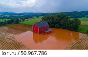A striking red barn submerged in muddy floodwaters amidst green farmland, depicting the severe impact of a natural disaster for climate change topics. Стоковое AI-изображение, AI-художник Данила Большаков / Фотобанк Лори