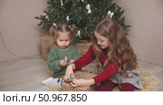 Two little girls, an older sister and a toddler, sit on the floor under the Christmas tree at home, playing with gifts and toys. Happy family and joy of winter holidays celebration concept. Footage. Стоковое видео, видеограф Ирина Михайличенко / Фотобанк Лори