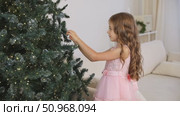 Side view cute little child girl in pink princess dress is decorating Christmas tree in living room at home. Merry Christmas and Happy New year winter holidays. High quality footage. Стоковое видео, видеограф Ирина Михайличенко / Фотобанк Лори