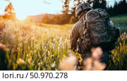 A military soldier, seen from the back with a large tactical backpack and helmet, walks through a sun-drenched field of wildflowers at golden hour, evoking a sense of journey and solitude. Стоковое AI-изображение, AI-художник Данила Большаков / Фотобанк Лори