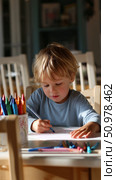 Young child draws with crayons while seated at a table in a bright room during daylight hours. Стоковое AI-изображение, AI-художник Сергей Толмачёв / Фотобанк Лори