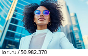 Confident businesswoman in a white blazer and blue sunglasses against a modern cityscape background. Стоковое AI-изображение, AI-художник Данила Большаков / Фотобанк Лори