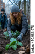 Focused young woman with red hair and grey beanie wearing gloves carefully plants green seedlings in rich dark soil, contributing to urban garden project with another person nearby. Стоковое AI-изображение, AI-художник Данила Большаков / Фотобанк Лори