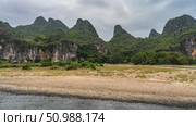 Green grass and bushes grow on the pebbly riverbank. Picturesque mountains against the sky and clouds. China. Li River. Li Jiang. Guilin. Стоковое фото, фотограф Вера Ивановна Кубарева / Фотобанк Лори