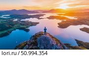 Majestic Sunset Panorama: Lone Figure on Mountaintop Overlooking Serene Fjord. Стоковое AI-изображение, AI-художник Данила Большаков / Фотобанк Лори