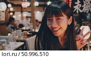 Young woman with black hair gesturing peace sign, winking playfully near traditional Japanese restaurant lanterns and kanji signage. Стоковое AI-видео, аниматор Данил Руденко / Фотобанк Лори