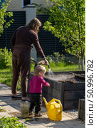 Family garden: a toddler learns about gardening while her mother waters the plants in a raised garden bed on a bright sunny day. Стоковое фото, фотограф Данила Большаков / Фотобанк Лори