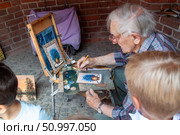 Elderly painter creating art outdoors, depicting a flower vase picture, surrounded by onlookers in an informal setting, Варшава, Польша (2018 год). Редакционное фото, фотограф Данила Большаков / Фотобанк Лори
