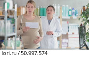Focused pregnant woman with prescription in hands looking around pharmacy trying to find right item while female pharmacist busy shelving supplies in background. Стоковое видео, видеограф Яков Филимонов / Фотобанк Лори