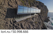 Luxurious, modern house is built into the side of a cliff, overlooking the vast ocean, its large windows reflecting the serene blue seascape and offering breathtaking views. Стоковое AI-видео, аниматор Данил Руденко / Фотобанк Лори