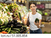 In supermarkets vegetable store, female customer takes some cucumber from display shelf. Woman selects necessary vegetables. Стоковое фото, фотограф Яков Филимонов / Фотобанк Лори