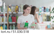 Girl in summer clothes holds dishwashing detergent in her hands, standing in a supermarket against the background of other visitors. Customer chooses a gel for washing dirty dishes. Стоковое видео, видеограф Яков Филимонов / Фотобанк Лори