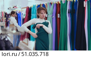Customer in store has removed hanger with floor-length gown from window and is considering product. Woman client imagines successful new summer outfit. Стоковое видео, видеограф Яков Филимонов / Фотобанк Лори