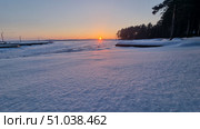 A winter sunset over a frozen lake. The sun is setting on the horizon, painting the snow in soft pink and gold hues. In the foreground is a smooth, shiny blanket of snow, sparkling in the evening light. On the left are the docks extending into the lake, and on the right is the dark silhouette of a pine forest. The atmosphere of peace and quiet, typical of a frosty winter evening. Стоковое фото, фотограф Анна Кривицкая / Фотобанк Лори