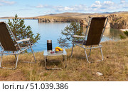 Rest on the shore of Baikal Lake concept. Thermos, fruits on camping table and folding chairs on coast of Olkhon Island on sunny summer day. Scenic landscape. Travels and outdoor recreation. Стоковое фото, фотограф Виктория Катьянова / Фотобанк Лори